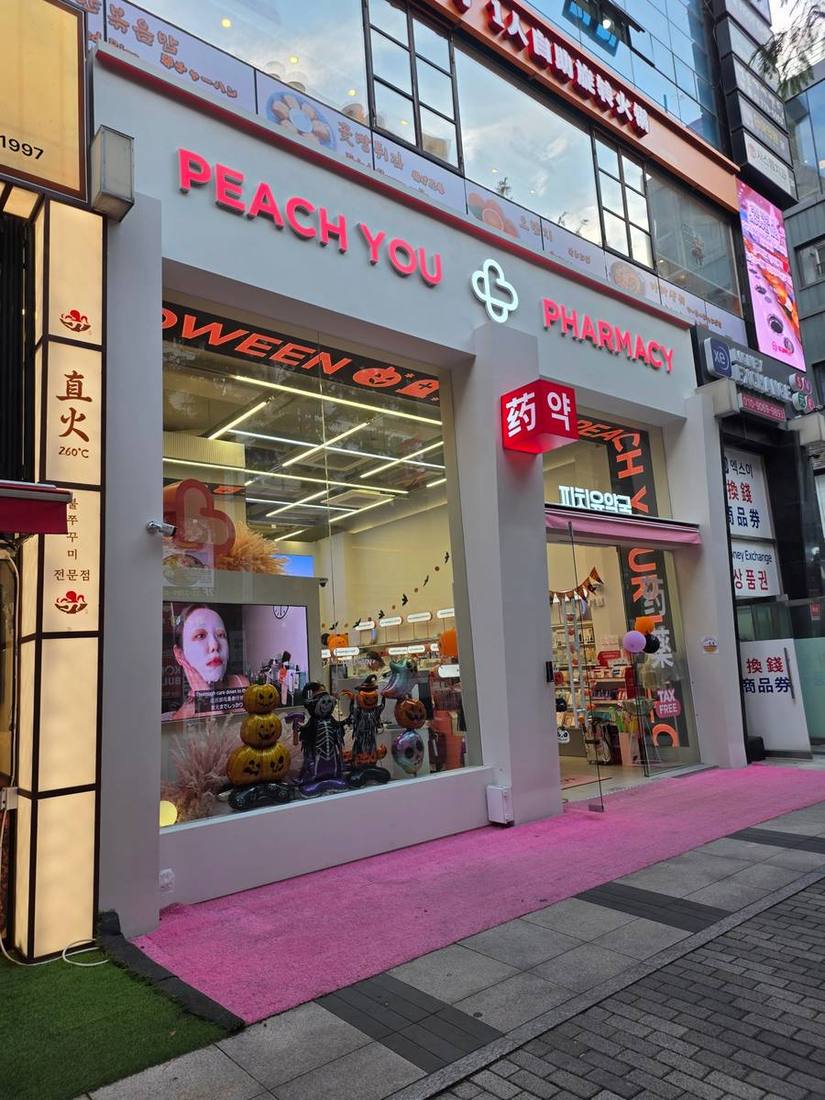 핼러윈 장식으로 꾸민 전면 통유리 쇼윈도와 핑크색 카펫이 눈에 띄는 피치유약국. 중국어로 ‘약’을 뜻하는 글자와 TAX FREE 안내가 보입니다. 오른쪽에는 환전소가 나란히 자리합니다.