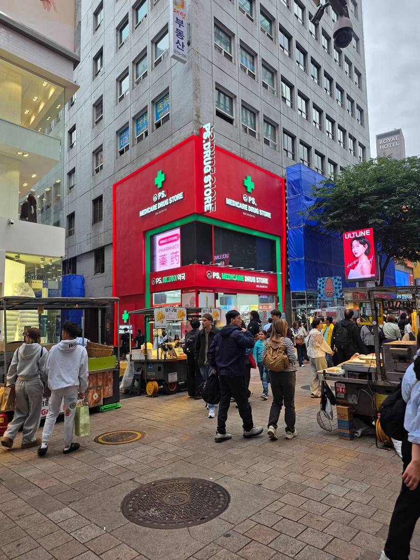 회색 타일 건물의 저층 코너를 붉은색 박스형 외관으로 감싼 약국. 초록색 십자와 다국어 약국 안내가 눈에 띕니다. 위층에는 치과와 의원이 자리하고, 옆 건물은 공사 중입니다.