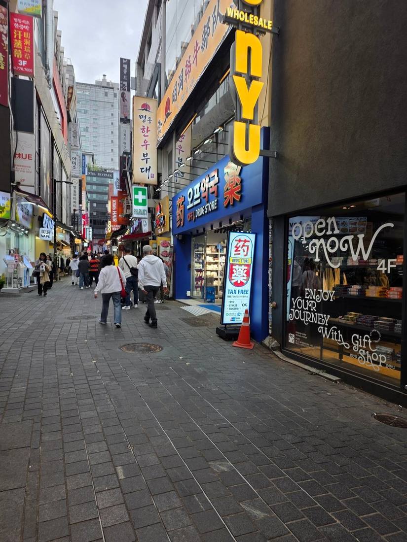 파란색 파사드의 오프약국. 입구에는 외국어 안내와 TAX FREE 표시, 주요 간편결제 로고가 적힌 입간판이 서 있습니다. 오른쪽에는 대형 액세서리 전문점 뉴뉴(NYU NYU)가 나란히 자리하고, 4층 매장을 안내하는 문구가 보입니다.