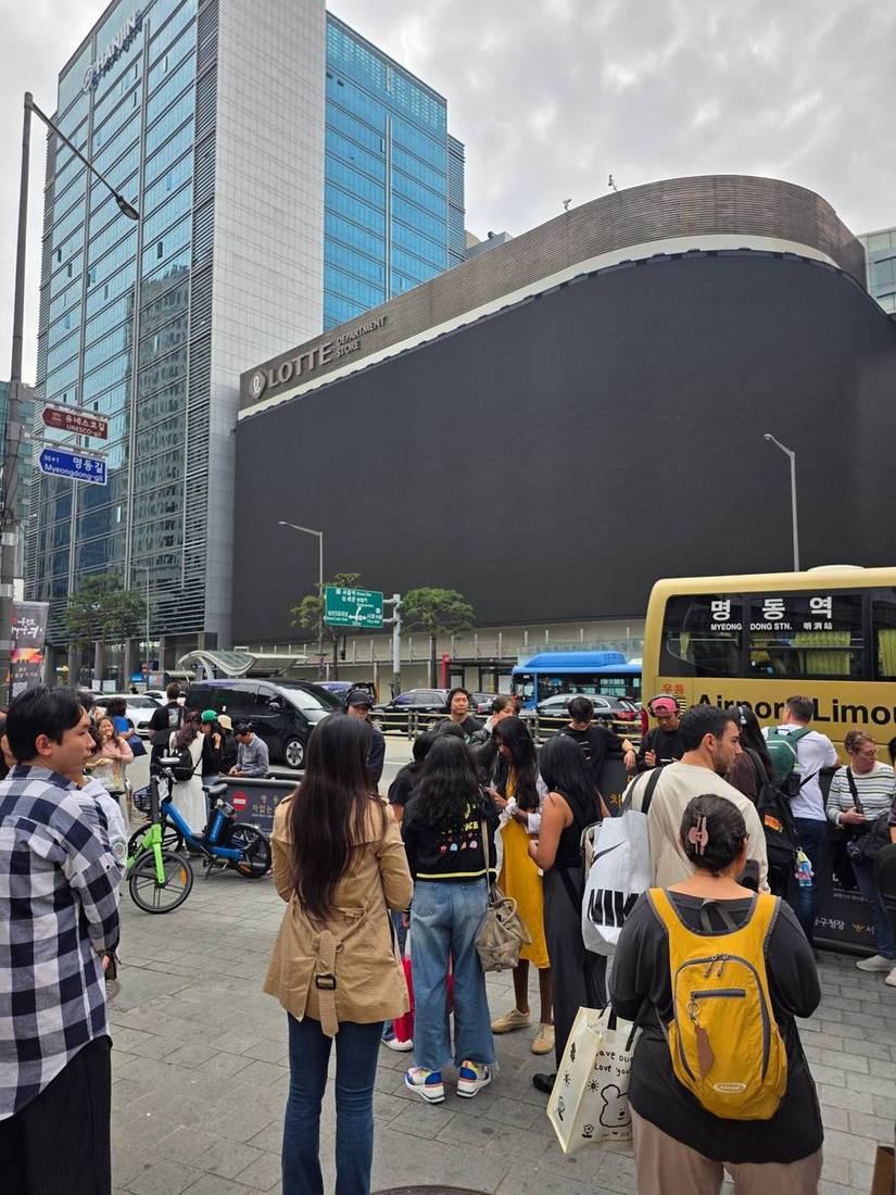롯데백화점 본점 앞 명동역 공항리무진 정류장. 노란 리무진 버스와 시내버스, 택시가 오가고 쇼핑백과 배낭을 멘 외국인 관광객 무리가 모여 있습니다.