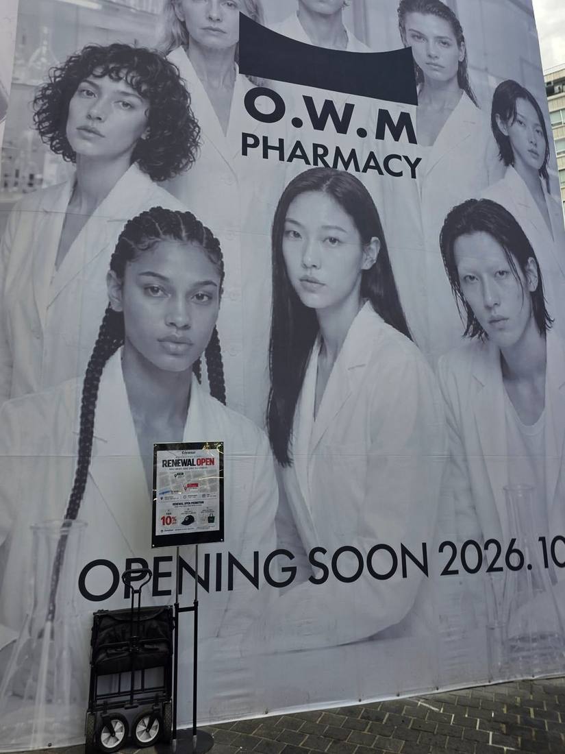 건물 전면을 덮은 흑백 대형 가림막. 흰 가운 차림 모델들 사진 위에 ‘O.W.M PHARMACY’와 ‘OPENING SOON 2026.10’ 문구가 크게 보입니다. 그 앞에는 패션 브랜드 커버낫의 ‘RENEWAL OPEN’ 이전 안내판이 서 있습니다.