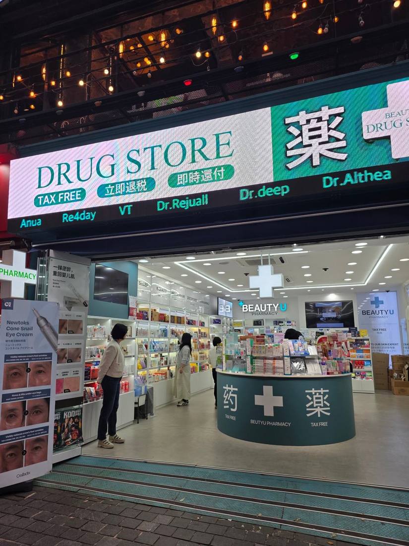 건물 전면을 가득 채운 LED 간판에 ‘DRUG STORE’와 중국어·일본어 즉시환급 안내가 표시되어 있습니다. K-뷰티 브랜드도 간판에 함께 소개되고, 매장 중앙에는 중국어와 일본어로 ‘약’을 뜻하는 글자가 새겨진 둥근 카운터가 있습니다.